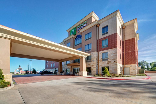 Four-story hotel building with a portico entrance. Brick and stone exterior, blue sky background, and parked cars near the entrance.