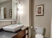 Modern bathroom with a square sink on a granite countertop, a wall mirror, a toilet, and a framed picture. Bright lighting fixture next to the mirror. Towels and toiletries visible.