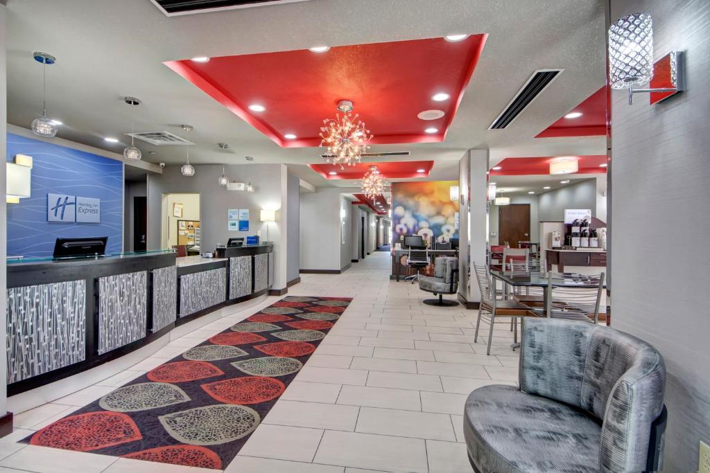 Hotel lobby with modern decor, featuring red ceiling panels, pendant lights, checkered seating, a reception desk on the left, and a patterned rug on the tile floor.