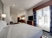 Hotel room with a large bed, flat-screen TV, and kitchenette. Natural light streams through a window with blue curtains, creating a bright and airy atmosphere.