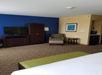 Hotel room with blue and beige walls, a TV on a dresser, an armchair, a small table, a wardrobe, and a kitchenette area in the corner.