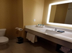 Hotel bathroom with a toilet, sink, illuminated mirror, towels, toiletries, and a small trash bin. Beige wallpaper and tiled floor.