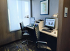 Small office space with two workstations, each with a computer, keyboard, and lamp, separated by a central printer. Black chairs face the desks, which have a window and decorative art above.