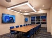 A modern conference room with a long wooden table, blue chairs, two wall-mounted screens, and large windows.