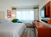 A hotel room with a king bed, a large window letting sun in, a green accent chair, a desk, a dresser, and a TV mounted to the wall.