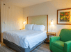 A hotel room with a white king bed, two night stands with lamps, a green accent chair, and a painting hanging on the wall.