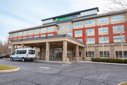 The tan stone and red exterior of the Holiday Inn Express Columbus Airport – Easton with the hotel shuttle parked in front.