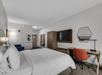 A modern hotel room with a large bed, wall-mounted TV, orange desk chair, hexagonal mirror, and wall art.