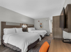 Hotel room with two double beds, a wall-mounted TV, a wooden desk with an orange chair, and a modern abstract painting on the wall.