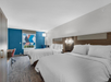 Hotel room with two double beds, modern decor, a blue accent wall, desk with chair, window with abstract art blinds, and neutral color palette.