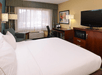 Hotel room with a king-sized bed, TV on a dresser, desk and chair, armchair, and window with curtains. The room has neutral tones and a modern style.