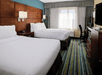 Hotel room with two double beds, striped carpet, wooden furniture, a desk with chair, and a TV. Large window with curtains allows natural light. Walls are painted teal above the bed.