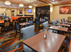 Restaurant dining area with wooden tables, assorted chairs, and colorful carpet. Artwork and TV on walls, pendant lights overhead, and fireplace in the background.