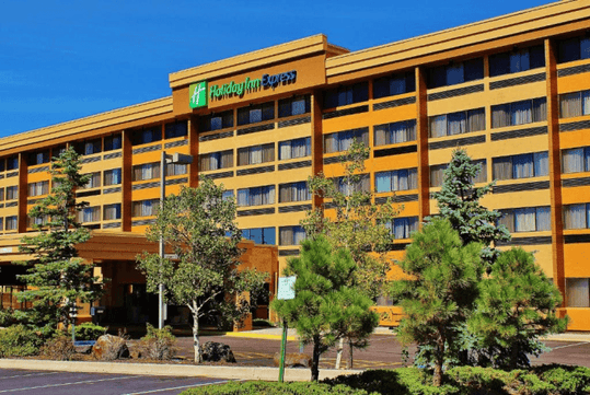 Exterior view of a Holiday Inn Express hotel with multiple floors, large windows, and trees in the foreground under a clear blue sky.