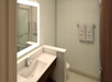 A modern bathroom with a sleek vanity, illuminated mirror, and a wall-mounted towel rack with two towels. The room has light-colored textured walls and a partially visible shower area.