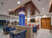 Modern lounge area with blue and red chairs, wooden tables, and a central column. Bright lighting and a large wall display create an inviting atmosphere.