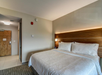 Hotel room with a large bed, wooden headboard, white linens, and wall lighting. The room has neutral walls, a carpeted floor, and a wooden door leading to a hallway.