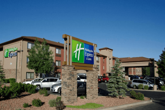 Hotel exterior with a "Holiday Inn Express & Suites" sign in the foreground. Several cars are parked in the lot under a clear blue sky.