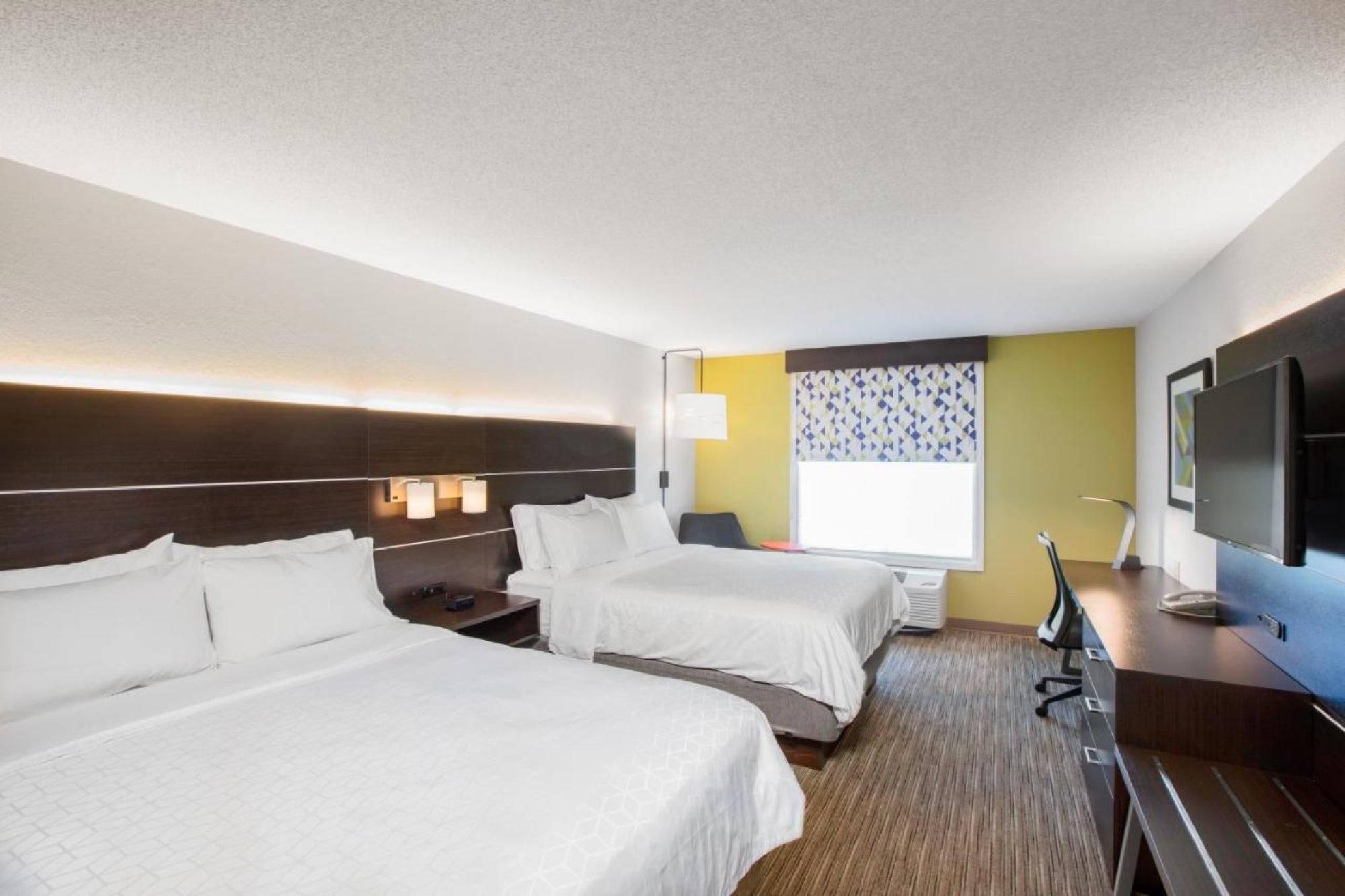 Hotel room with two neatly made beds, a desk with a chair, a flat-screen TV, and a window with patterned blinds.