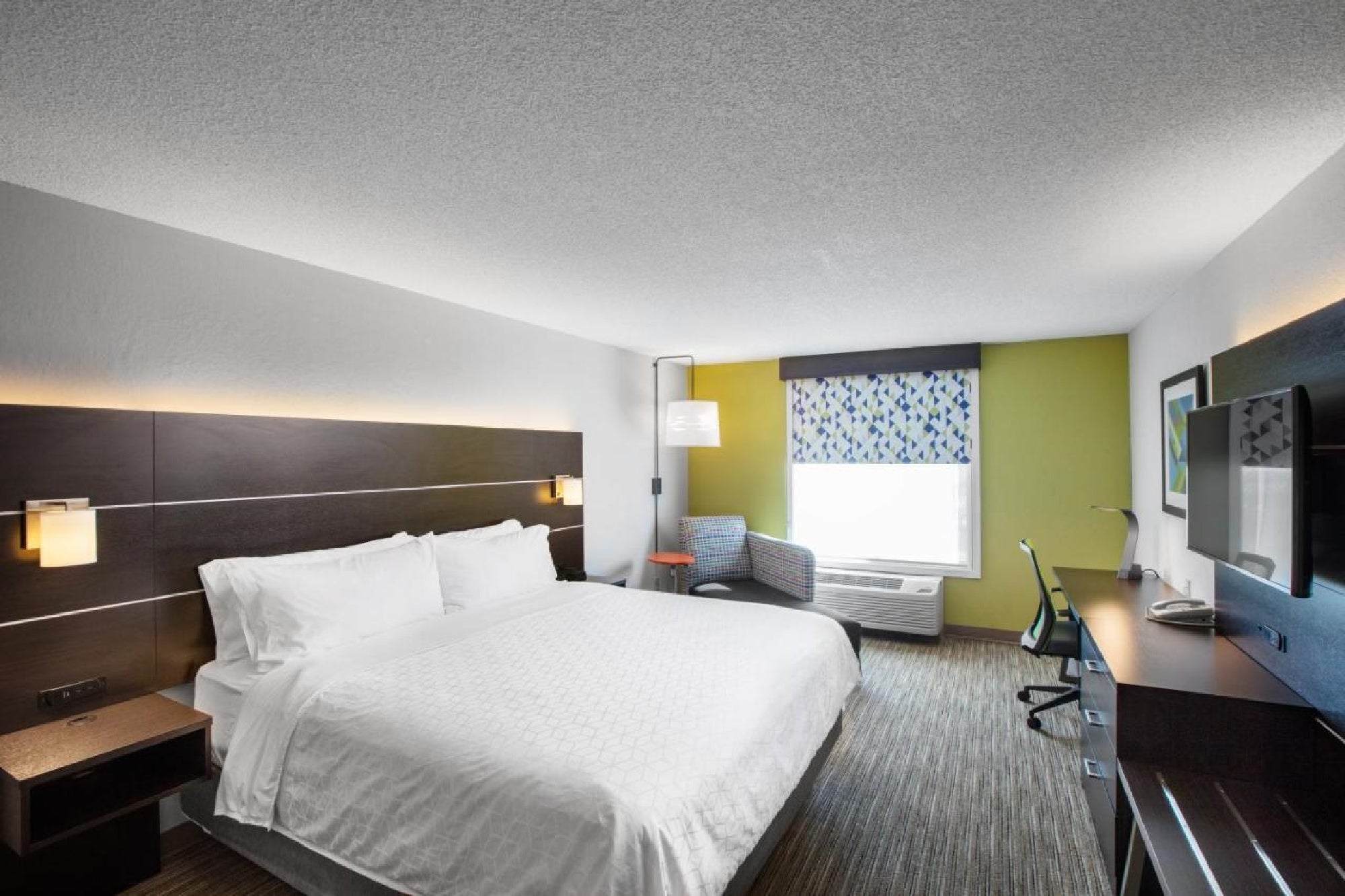 Modern hotel room with a large bed, wall-mounted TV, desk, chair, and window with patterned drapes. The decor features neutral tones with a splash of yellow on one wall.