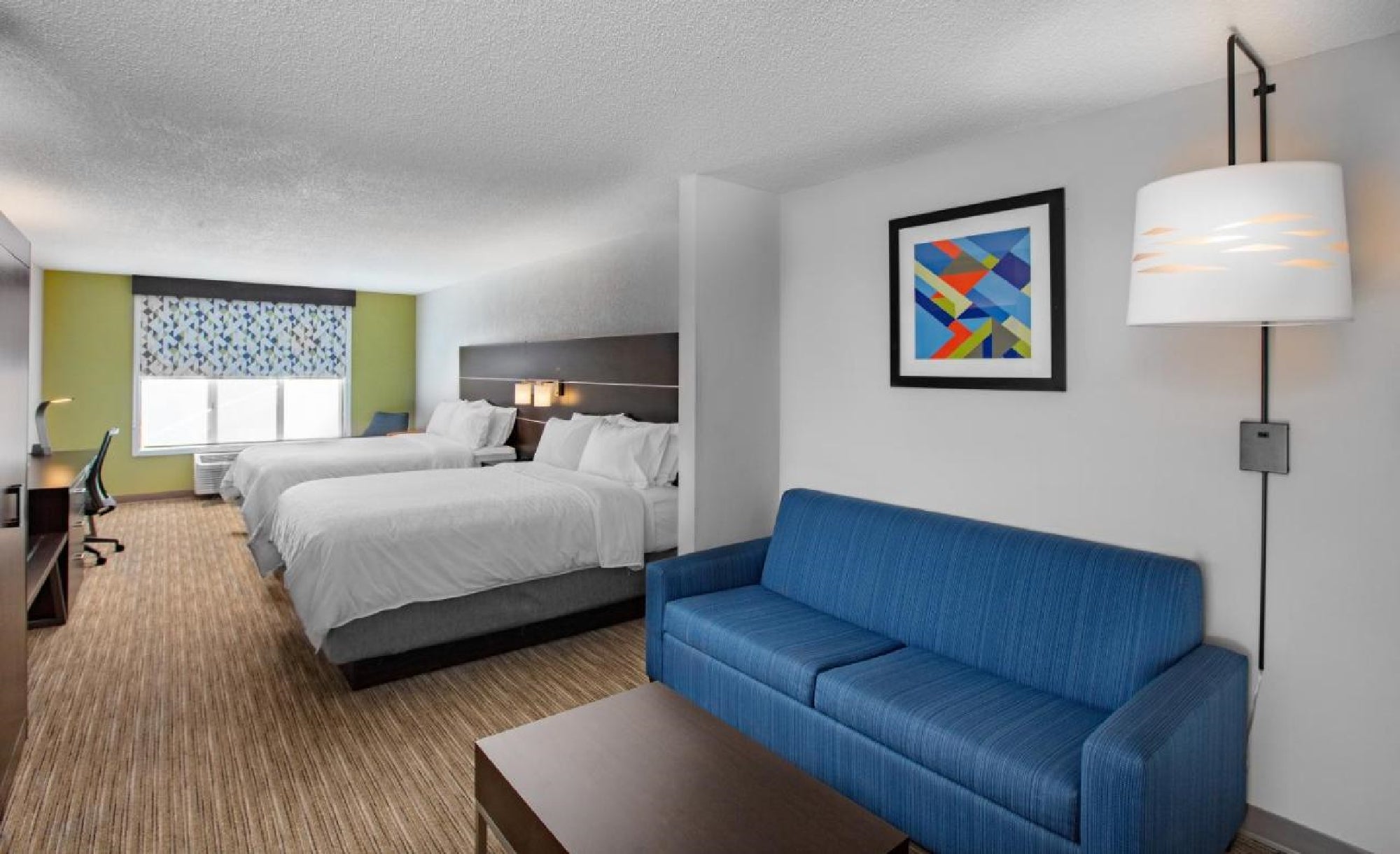 Hotel room with two double beds, a blue sofa, wall art, and a window. A lamp is mounted on the wall above the sofa.