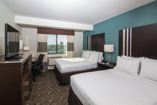 Hotel room with two beds, a TV, desk, and chair. A window with curtains shows a view of trees outside. The room has a teal accent wall.