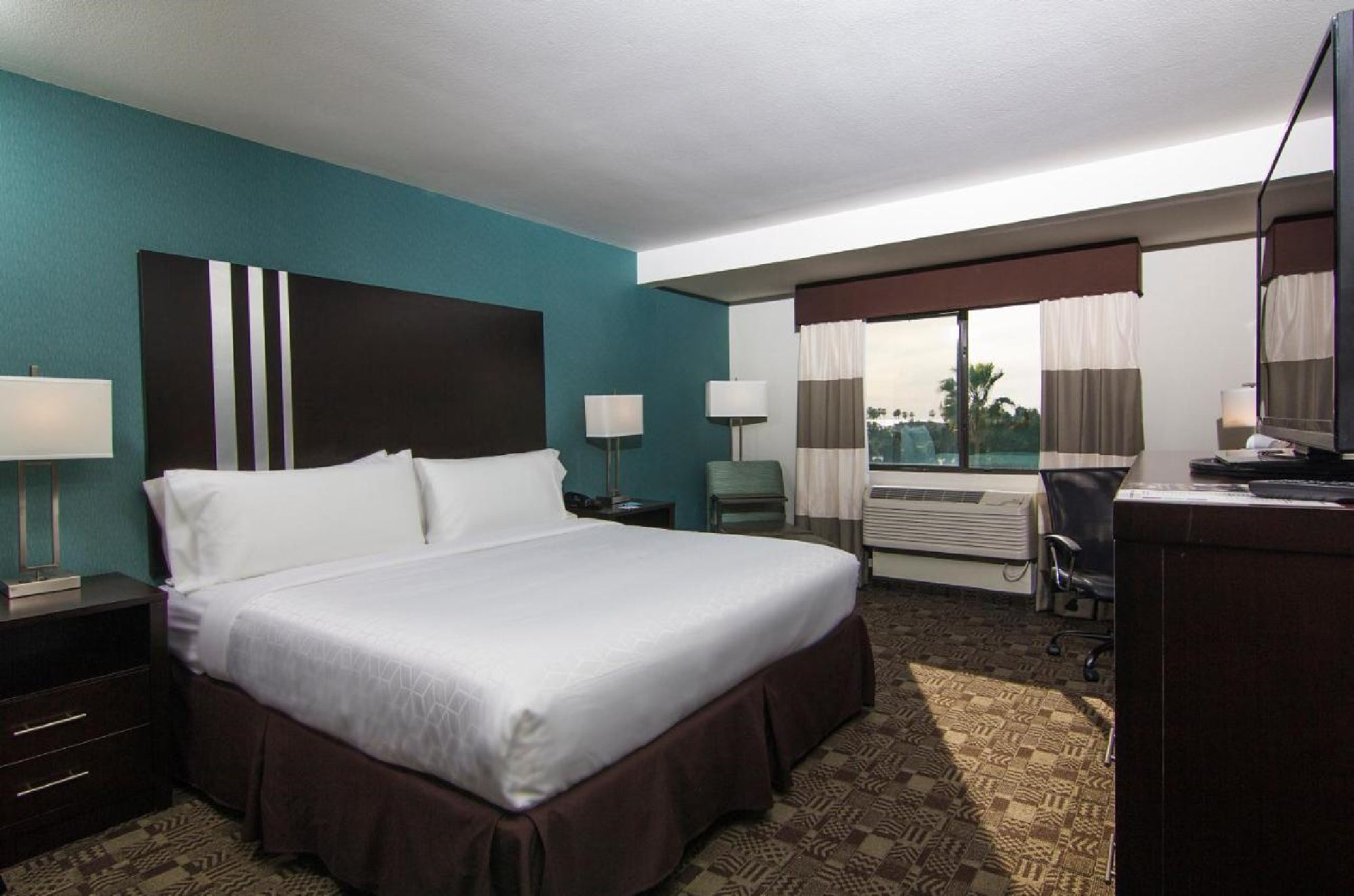 Hotel room with a king-sized bed, dark wood furniture, teal accent wall, large window, desk, chair, and lamps.