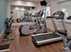 A gym room with treadmills, an elliptical machine, dumbbells, and a bench on a wooden floor, with mirrors and windows on the walls.