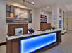 Hotel reception desk with a backlit panel, brochures, card machine, and display shelves with snacks and beverages in the background.