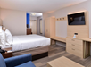 A hotel room with a bed, blue armchair, wall-mounted TV, dresser, and open closet. A small desk is visible near the entrance. The decor is modern and minimalistic.