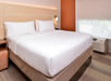 A hotel room featuring a large bed with white bedding, a wooden headboard, an orange accent wall, a side table, and a window with blinds.