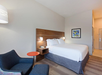 Hotel room with a large bed, wooden headboard, modern decor, navy armchair with a red side table, and a framed geometric artwork on a beige wall.
