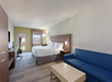 A hotel room with a king-sized bed, modern desk, blue sofa, coffee table, and wall-mounted TV. A large window and a green accent wall add natural light and color to the space.