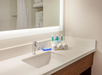 Bathroom countertop with a sink, a modern faucet, and a large illuminated mirror. On the counter are rolled towels and small bottles of toiletries.