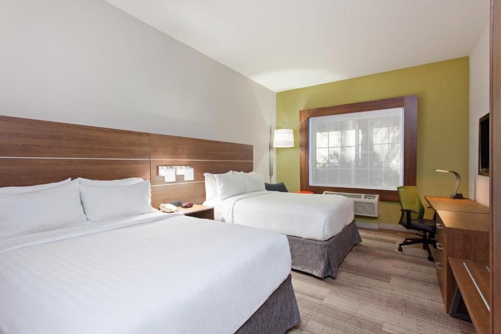 A hotel room featuring two double beds, a small desk with a chair, a window, and a wall-mounted TV.