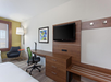 Hotel room with a modern desk setup, flat-screen TV mounted on a wooden panel, artwork on the wall, armchair by the window, and a bed in the foreground.