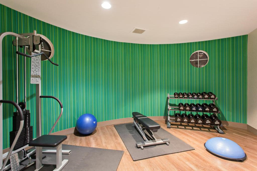 A gym room with a weight machine, bench, dumbbells, exercise balls, and a circular wall clock against a curved green striped wall.