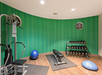 A gym room with a weight machine, bench, dumbbells, exercise balls, and a circular wall clock against a curved green striped wall.