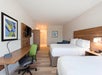 Hotel room with two double beds, a wooden desk with a green chair, a wall-mounted TV, and abstract artwork. The room has a carpeted floor and a small sofa in the corner.