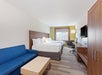 Hotel room with two double beds, blue sofa, wooden coffee table, desk with chair, wall-mounted TV, and a window with a view.