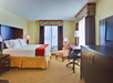 Hotel room with a large bed, two lamps, a desk with a chair, and a dresser. The room has two windows with curtains and striped valances. The walls are light yellow.