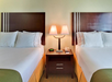 Hotel room with two double beds, each with white bedding and dark wooden headboards, separated by a nightstand holding a lamp, phone, and clock.