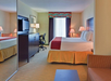 Hotel room with a king-size bed, a flat-screen TV on a dresser, a desk with a chair, a large window with curtains, and a wall mirror reflecting the room.