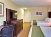 Hotel room with two double beds, a wall-mounted TV, desk with chair, coffee maker, and framed artwork. Walls are beige with carpeted floors in a similar tone. Door visible at the far end.