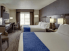 A hotel room with two double beds, each covered with a white duvet and blue runner. There’s a desk, chair, wall-mounted TV, and window with curtains. Warm lighting illuminates the room.