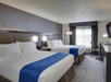 Hotel room with two large beds, white linens, and blue accent throws. A desk with a chair and TV is on the right. Natural light comes through a window with curtains.