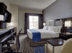 Modern hotel room with a king-sized bed, blue accents, a desk with a chair, a wall-mounted TV, and large window with curtains.
