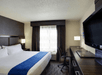 Hotel room with a king-sized bed, dark wood desk, chair, large TV, and a window with sheer curtains. Room is well-lit and modern in style.