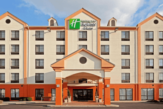 Exterior view of a Holiday Inn Express & Suites hotel with a prominent logo at the top, featuring multiple windows and an entrance under a covered walkway.