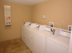A laundry room with four white washing machines and a wall-mounted dispenser for soap, bleach, and softeners. The floor is tiled, and the walls are beige.